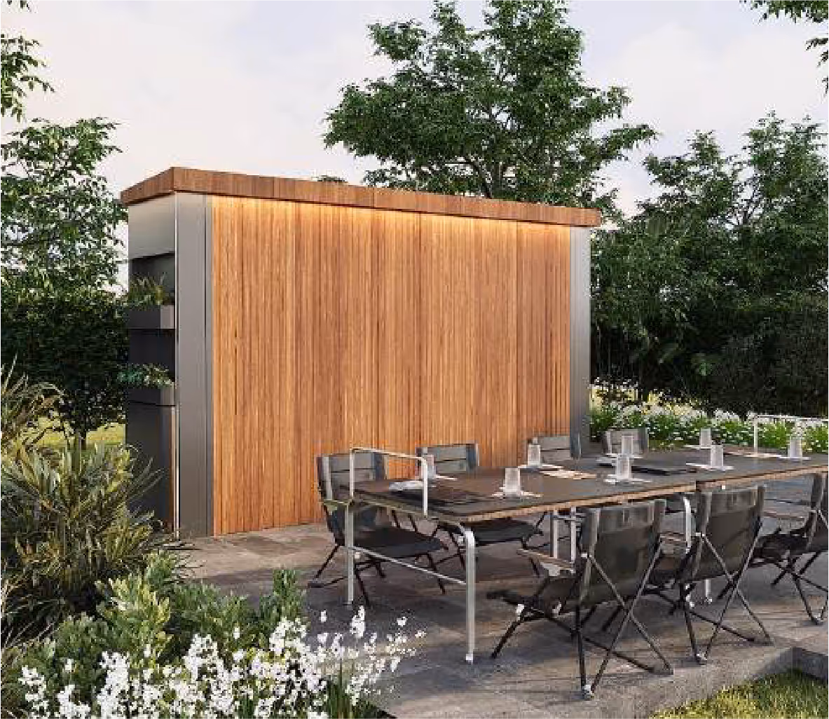 Outdoor dining area with a long table, black chairs, and a wooden privacy wall surrounded by lush greenery and white flowers.