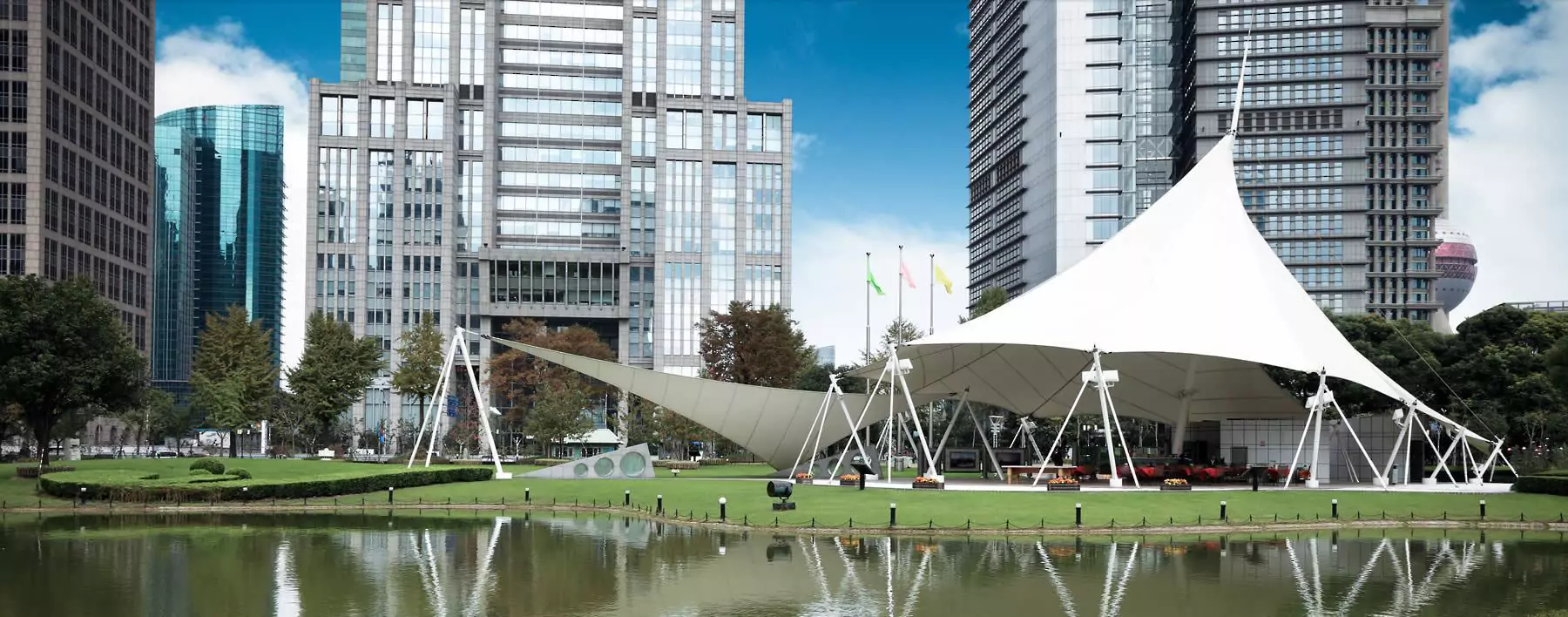 White tensile fabric canopy structure set in a green park with pond, surrounded by modern glass office buildings under a blue sky.