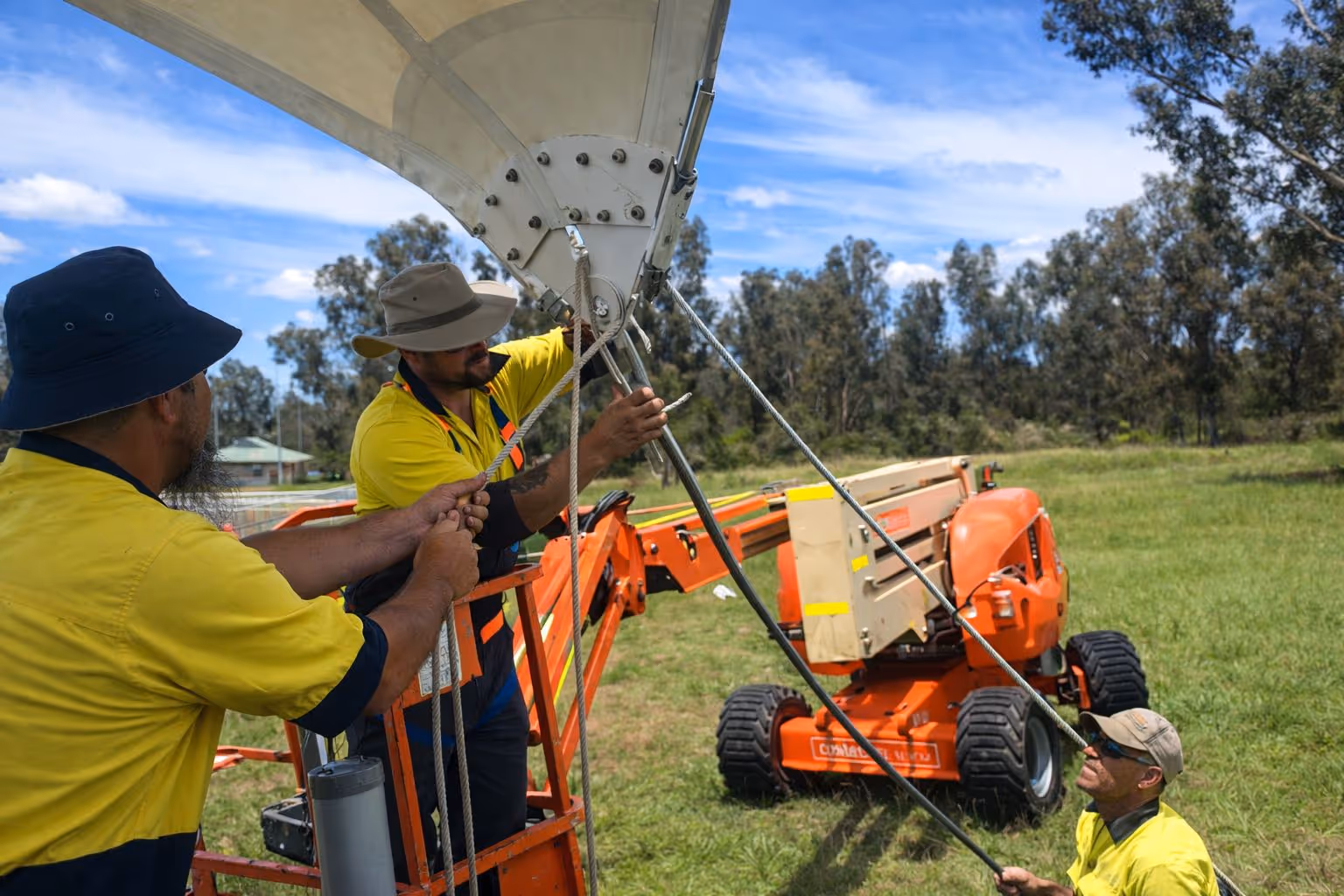 Three workers in yellow shirts and hats adjusting ropes on a large outdoor structure using an orange lift on a grassy field.