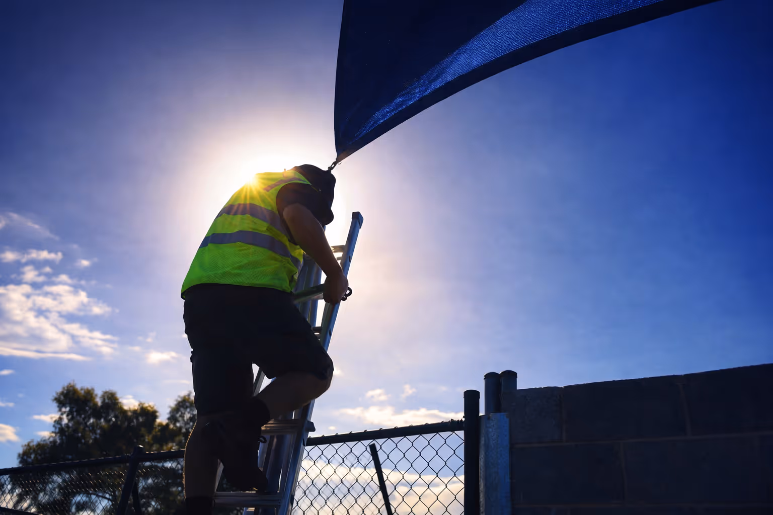 Worker wearing a neon safety vest climbing a ladder near a fence with the sun shining behind him.
