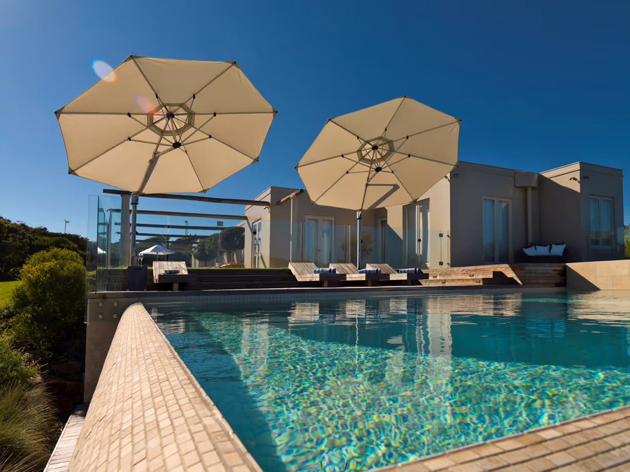 Modern outdoor pool with clear water, two large beige umbrellas, and wooden lounge chairs on a sunny day.